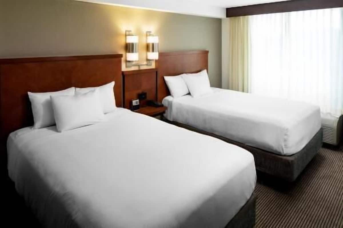 Dos camas de hotel juntas con sábanas blancas, cabecera de madera, mesita de noche con lámpara y ventana con cortinas.