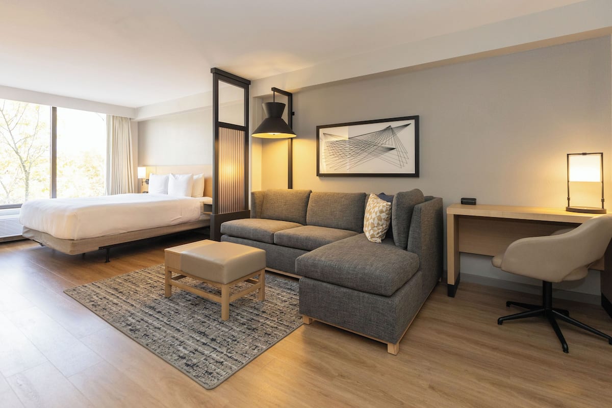 Una habitación de hotel moderna con una cama grande, un sofá seccional gris, un escritorio con una silla y un ventanal con vistas a árboles.