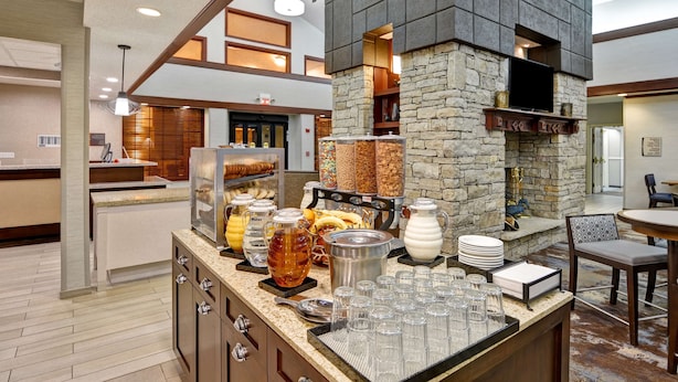 A breakfast bar with various food items and beverages, a stone fireplace, and a television mounted on the wall. - Homewood Suites Kansas City/Overland Park (Overland Park)