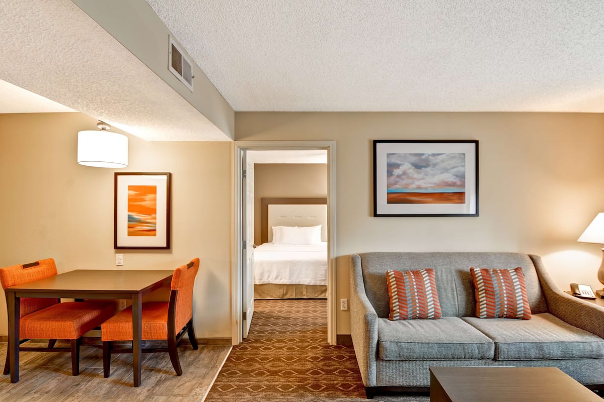 Habitación de hotel con una mesa de comedor, sillas anaranjadas, un sofá gris con cojines estampados, un cuadro en la pared y una cama al fondo.