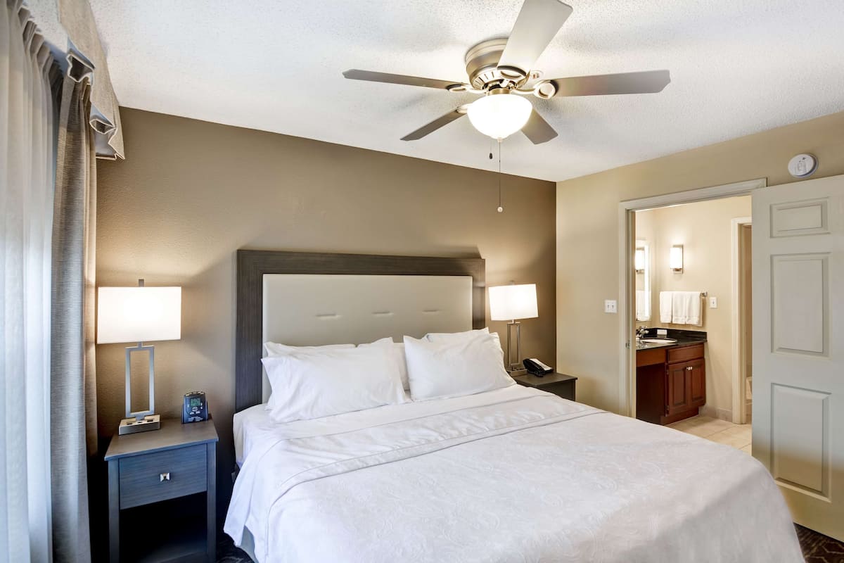 Una habitación de hotel con una cama, dos mesitas de noche con lámparas, un ventilador de techo, un baño con lavabo y espejo, y una puerta.