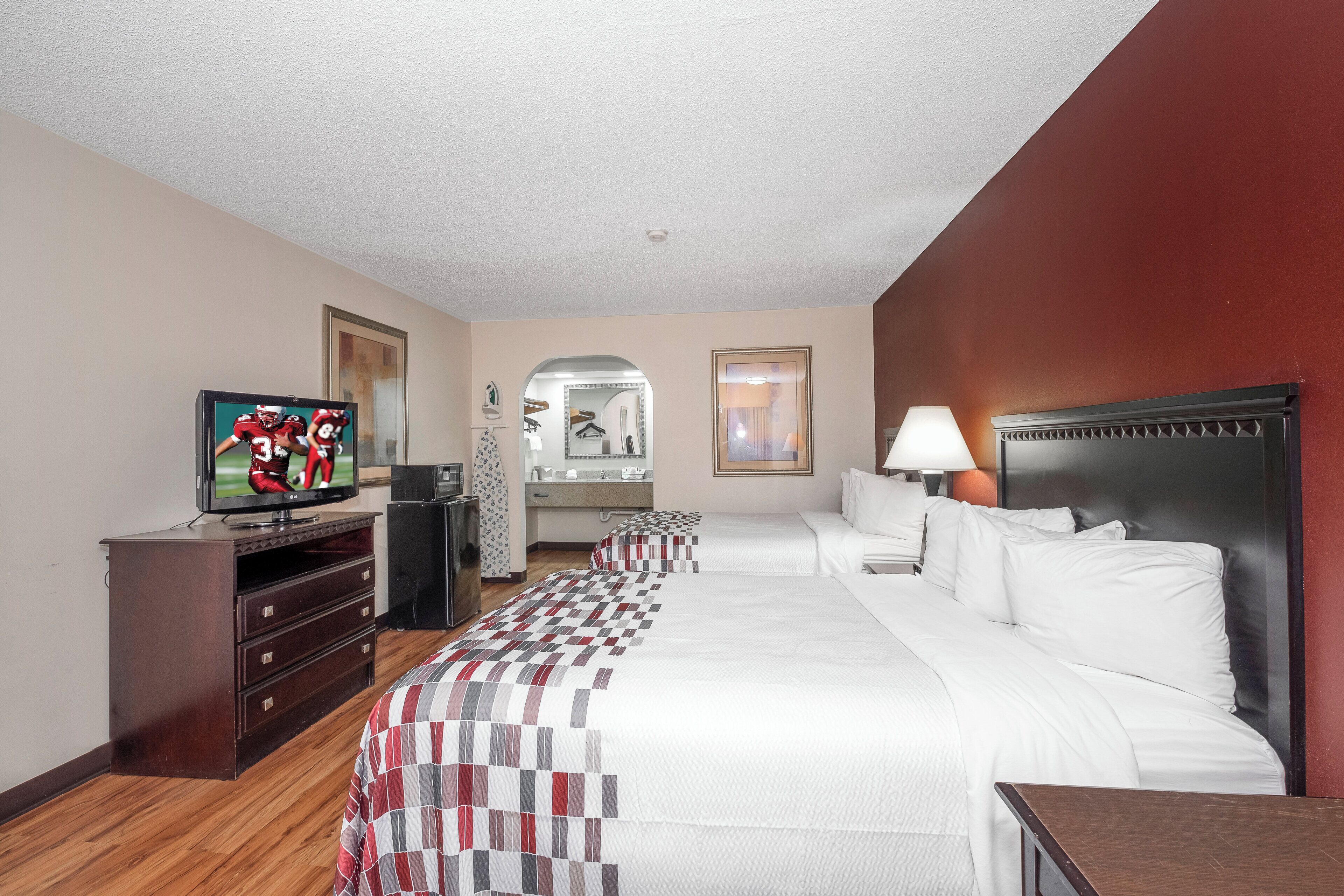 Ein Hotelzimmer mit einem großen Bett, einem Fernseher, einem Schminktisch, einem Spiegel und einem Nachttisch mit Lampe.