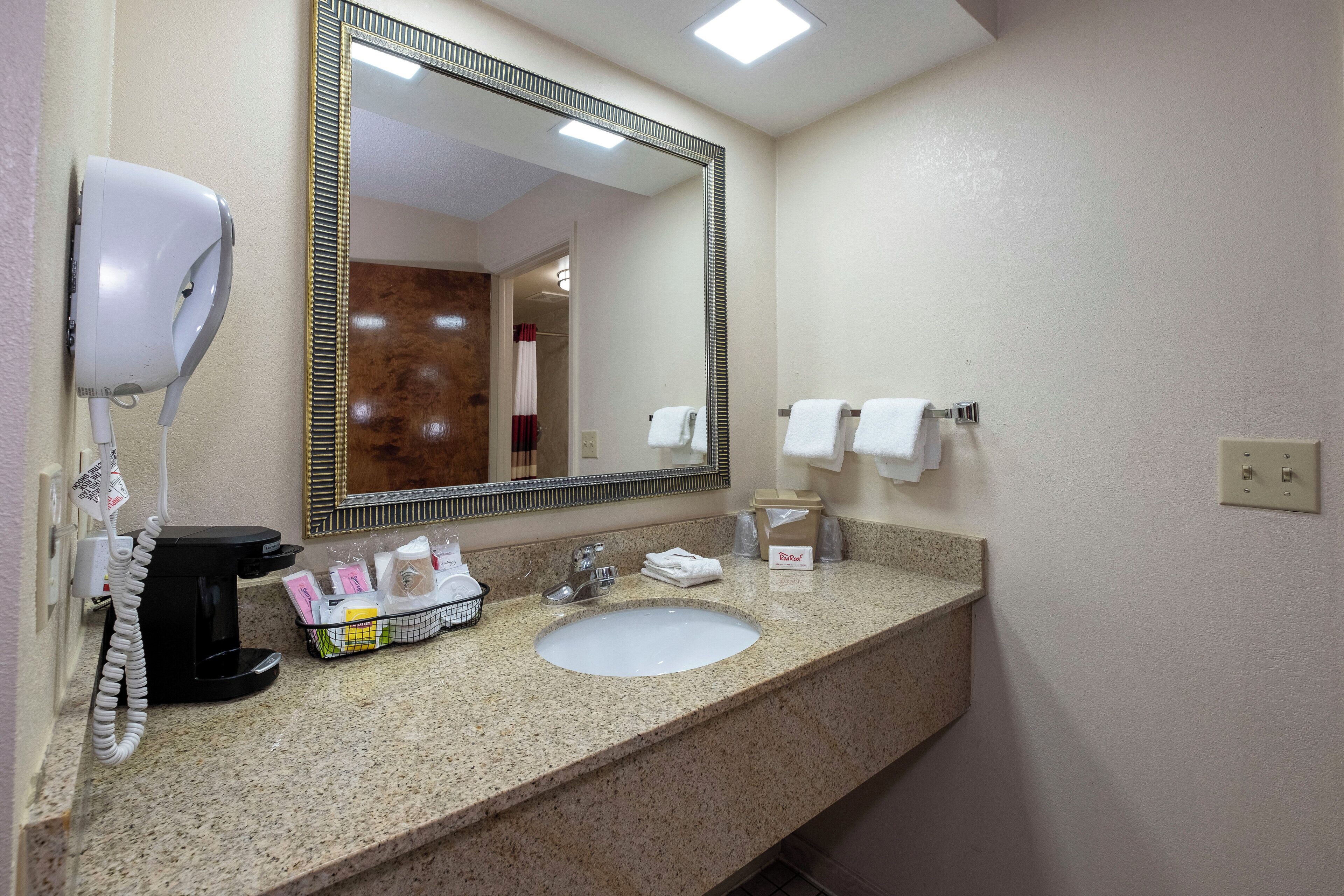 Une salle de bain d’hôtel équipée d’un comptoir en granit, d’un lavabo, d’un miroir, d’un sèche-cheveux, d’une machine à café et de produits de toilette.