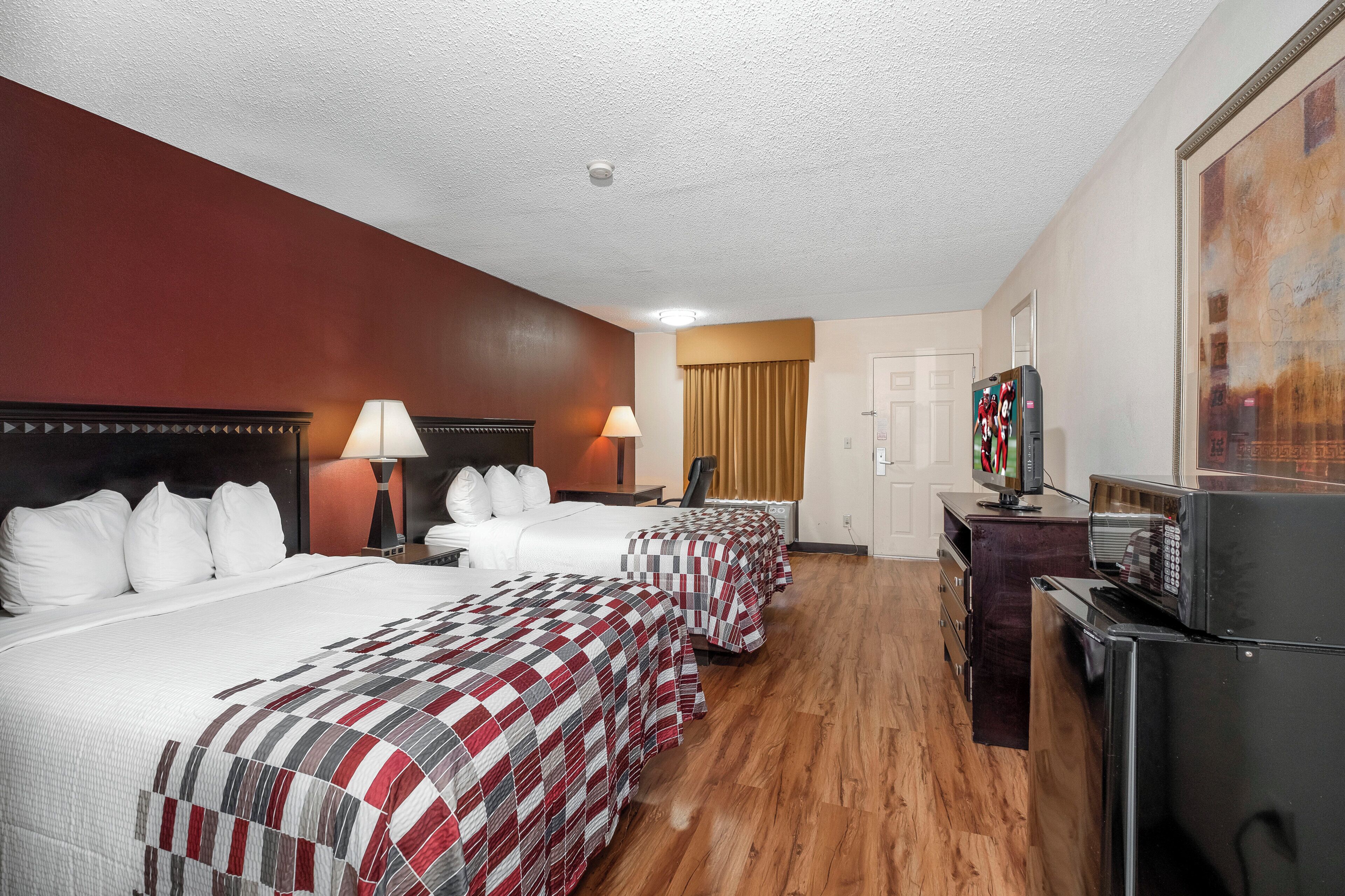 A hotel room with three beds, a television, a refrigerator, and a desk with a lamp.