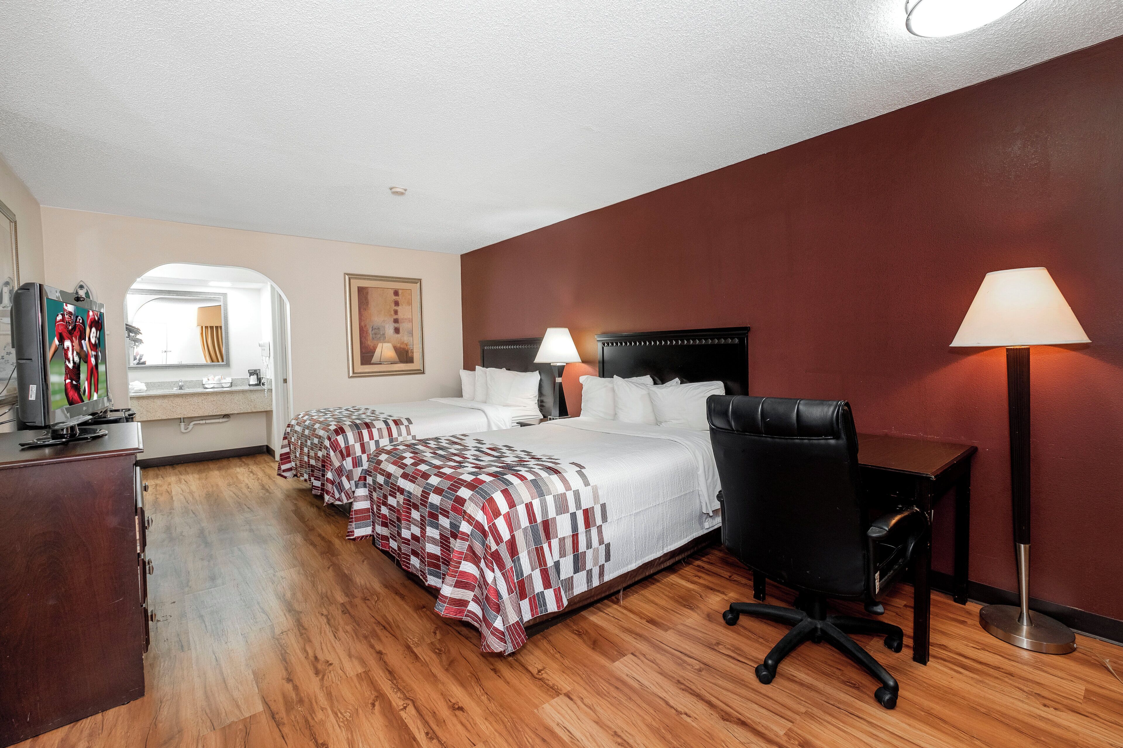 Quarto de hotel com cama, escrivaninha, cadeira, luminária e televisão sobre um aparador.