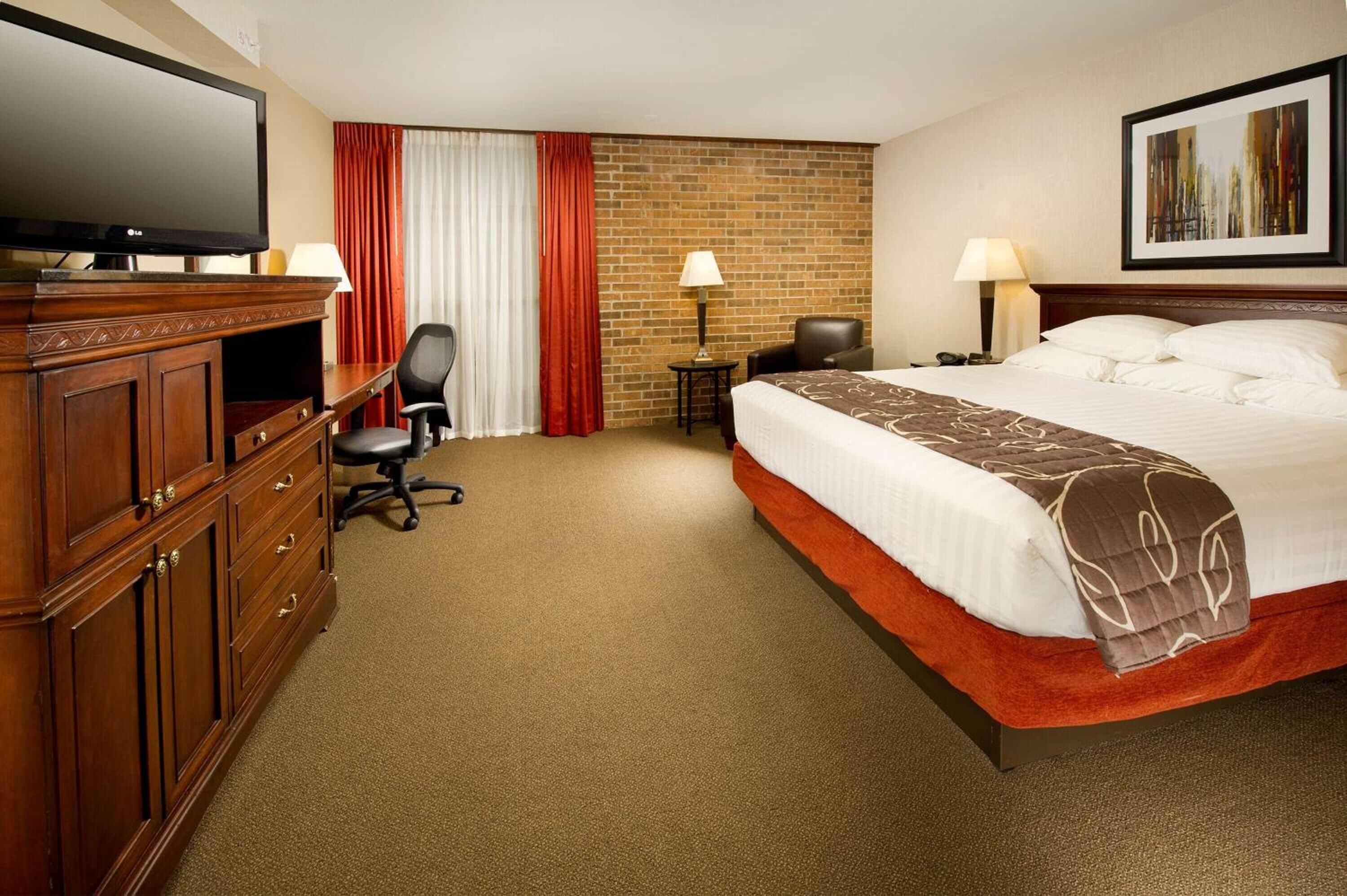 Une chambre d’hôtel avec un grand lit, une télévision, un bureau avec une chaise, une lampe et un mur en briques.