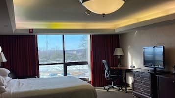 Habitación de hotel con cama, escritorio, silla, cómoda, televisor y un amplio ventanal con vistas al exterior.
