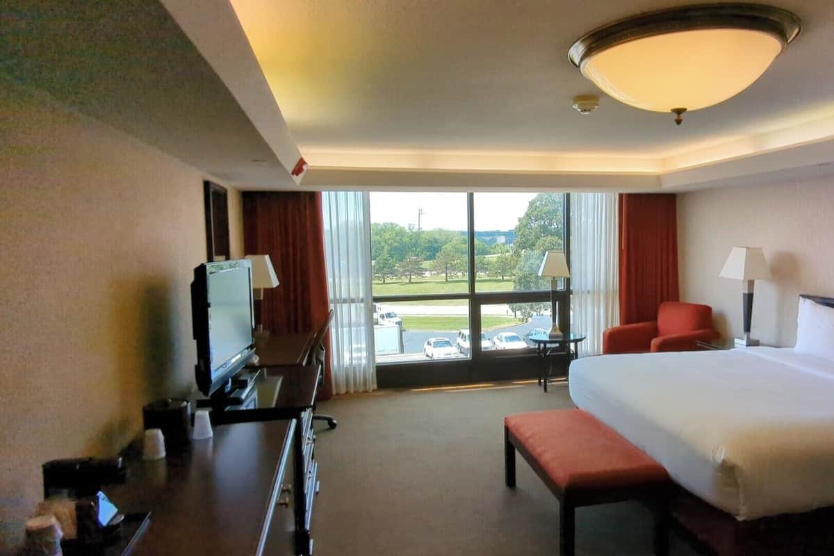 Una habitación de hotel con una cama grande, un escritorio con televisor, una silla y vistas a un estacionamiento y árboles.