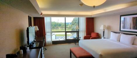 A hotel room with a large bed, a desk with a television, a chair, and a view of a parking lot and trees.