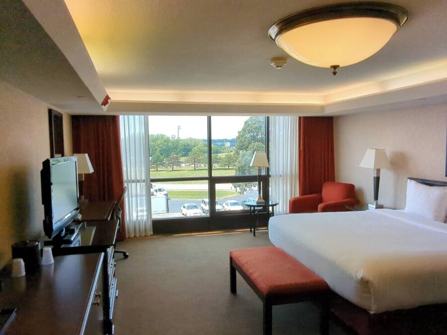 Een hotelkamer met een groot bed, een bureau met een televisie, een stoel en uitzicht op een parkeerplaats en bomen.