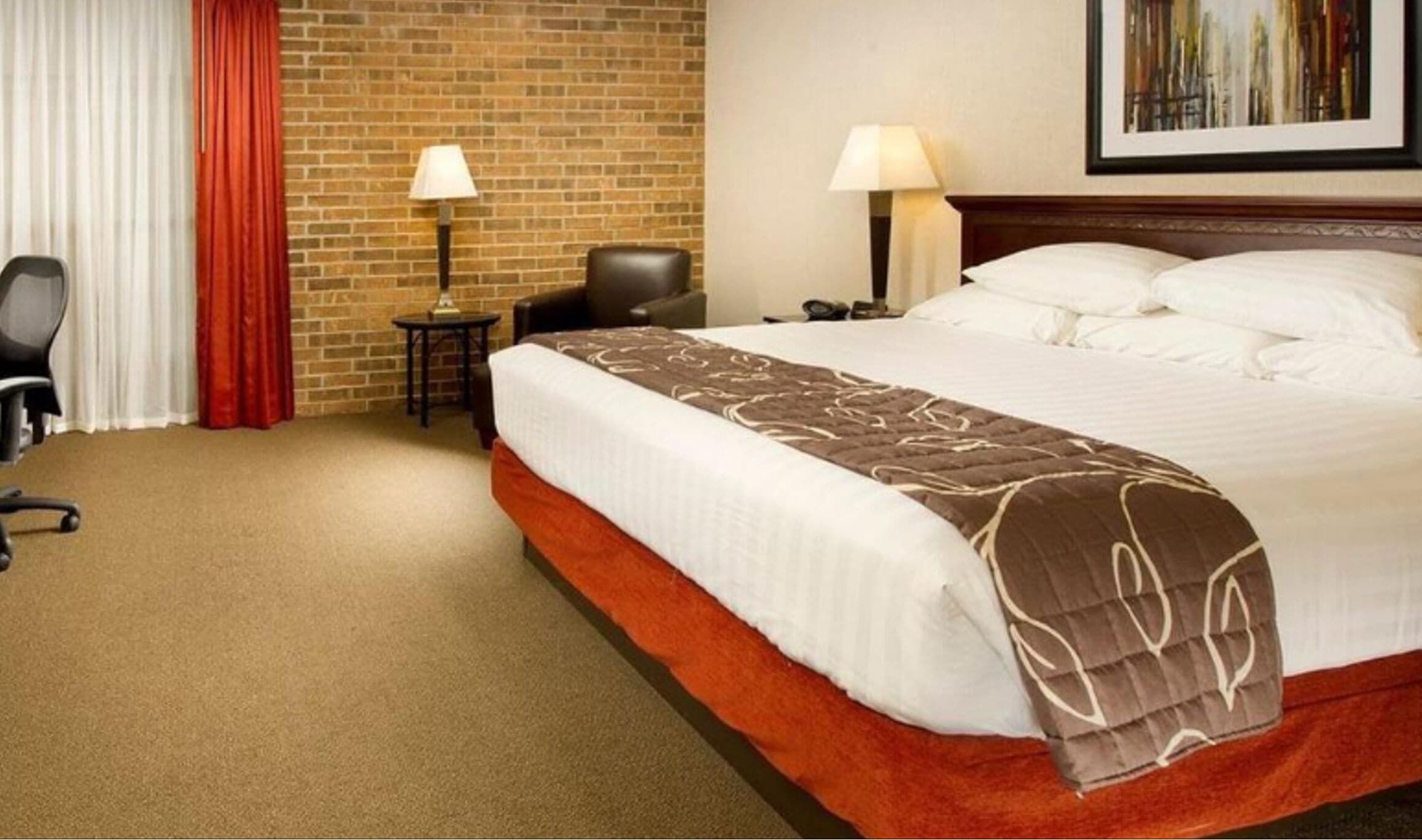 A hotel room with a large bed, a desk, a chair, a lamp, and a painting on the wall.