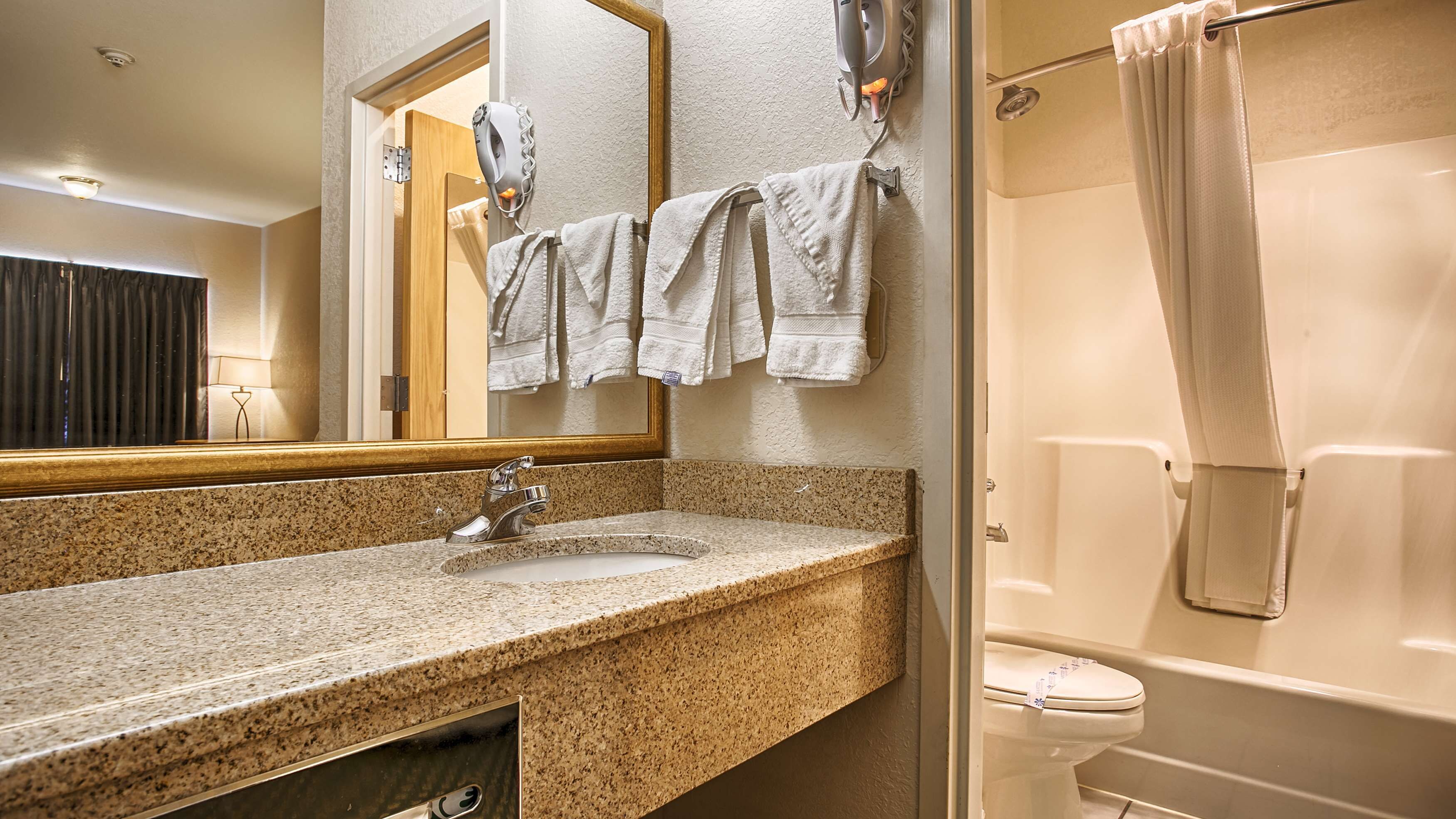A bathroom with a granite countertop sink, a mirror, a towel rack with multiple towels, a shower curtain, and a toilet.