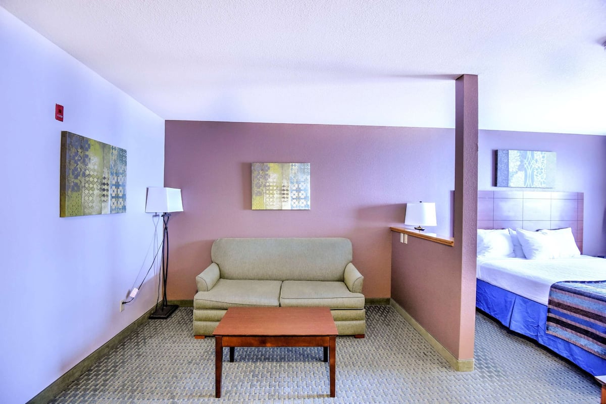 Ett hotellrum med en beige soffa, ett träbord, en säng med vita sängkläder och två väggmonterade lampor.