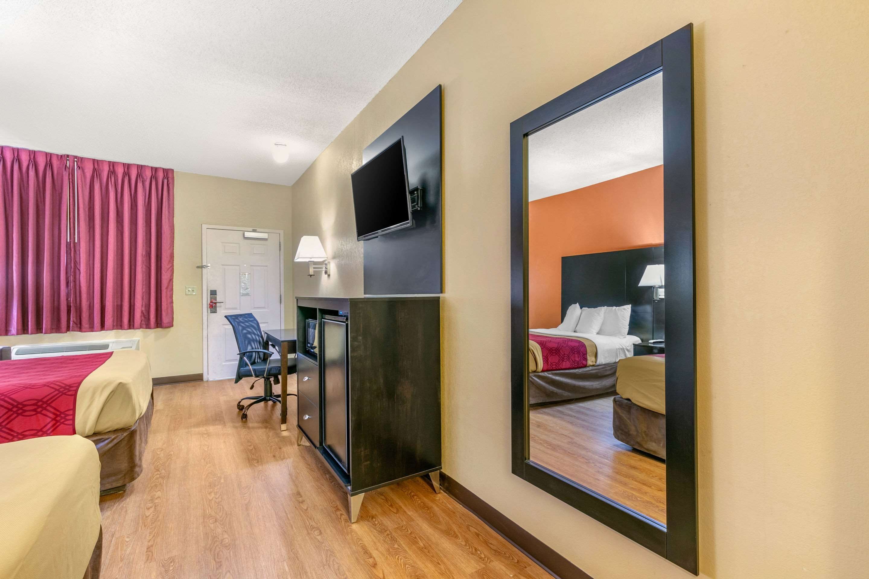 A hotel room with a bed, a desk with a chair, a TV mounted on the wall, and a large mirror reflecting the room.