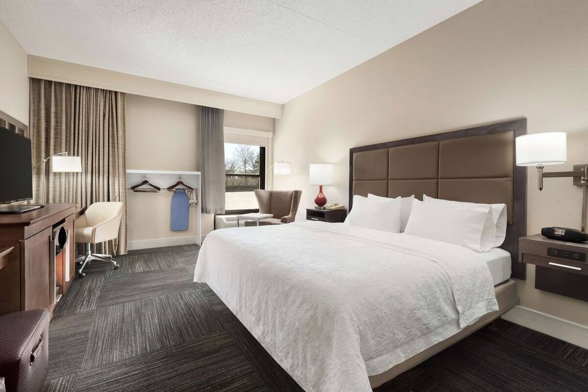 A hotel room with a large bed, a desk with a chair, a TV, a wardrobe, and a window with curtains.