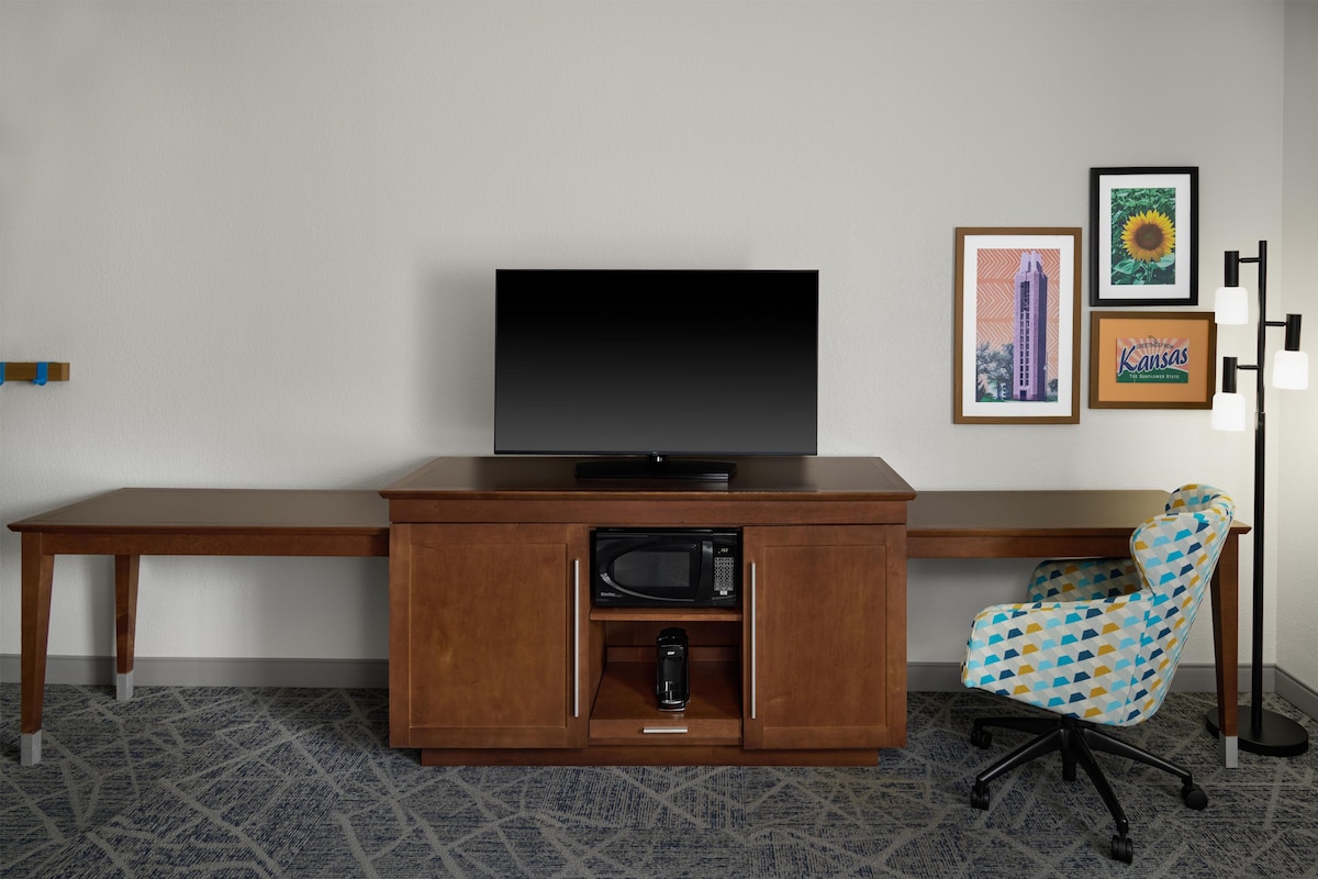 Une chambre d’h ôtel avec un bureau en bois, un téléviseur à écran plat, un four à micro-ondes, une machine à café et un fauteuil à motifs.
