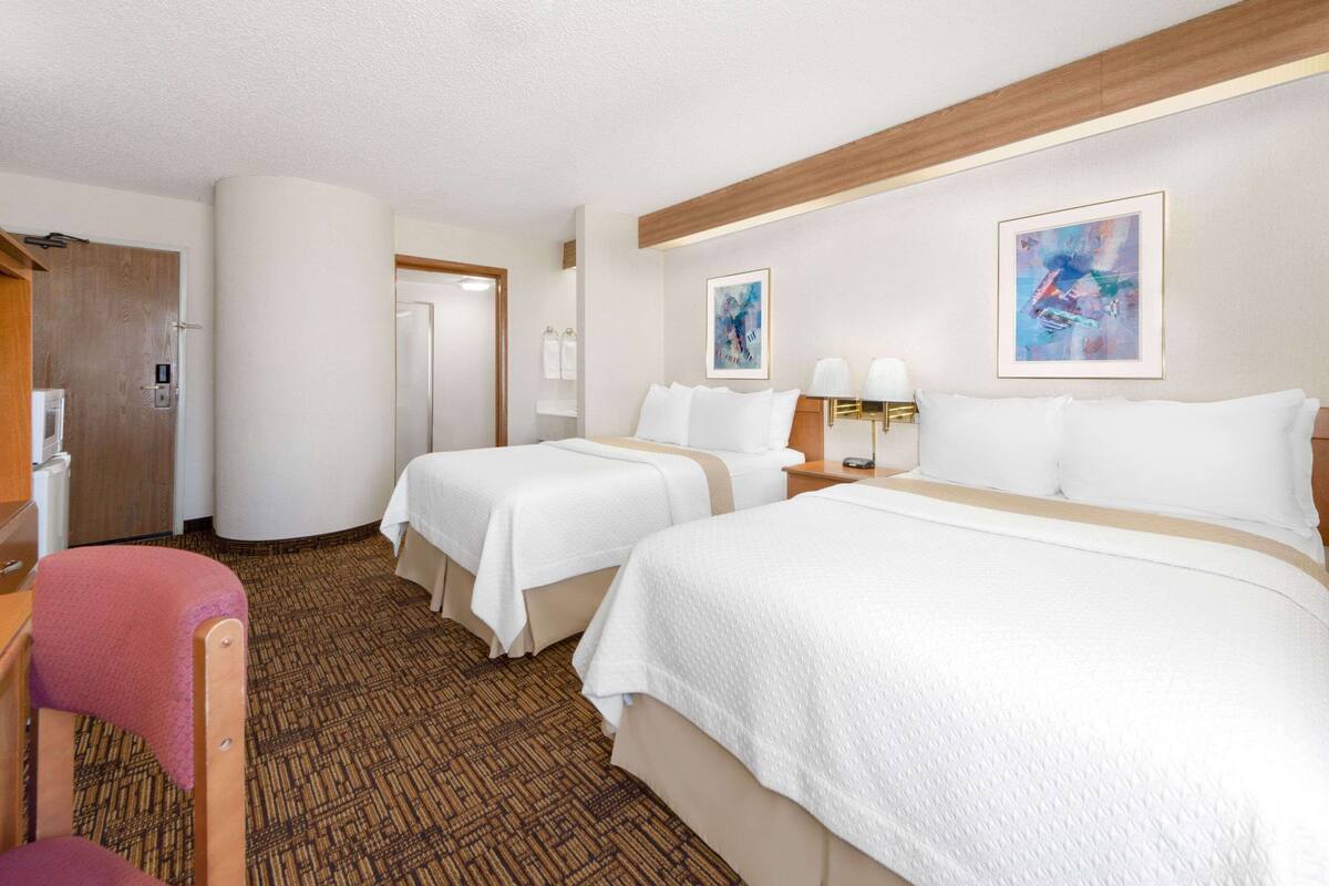 A hotel room with two beds, a wooden headboard, a desk with a chair, and a large window.