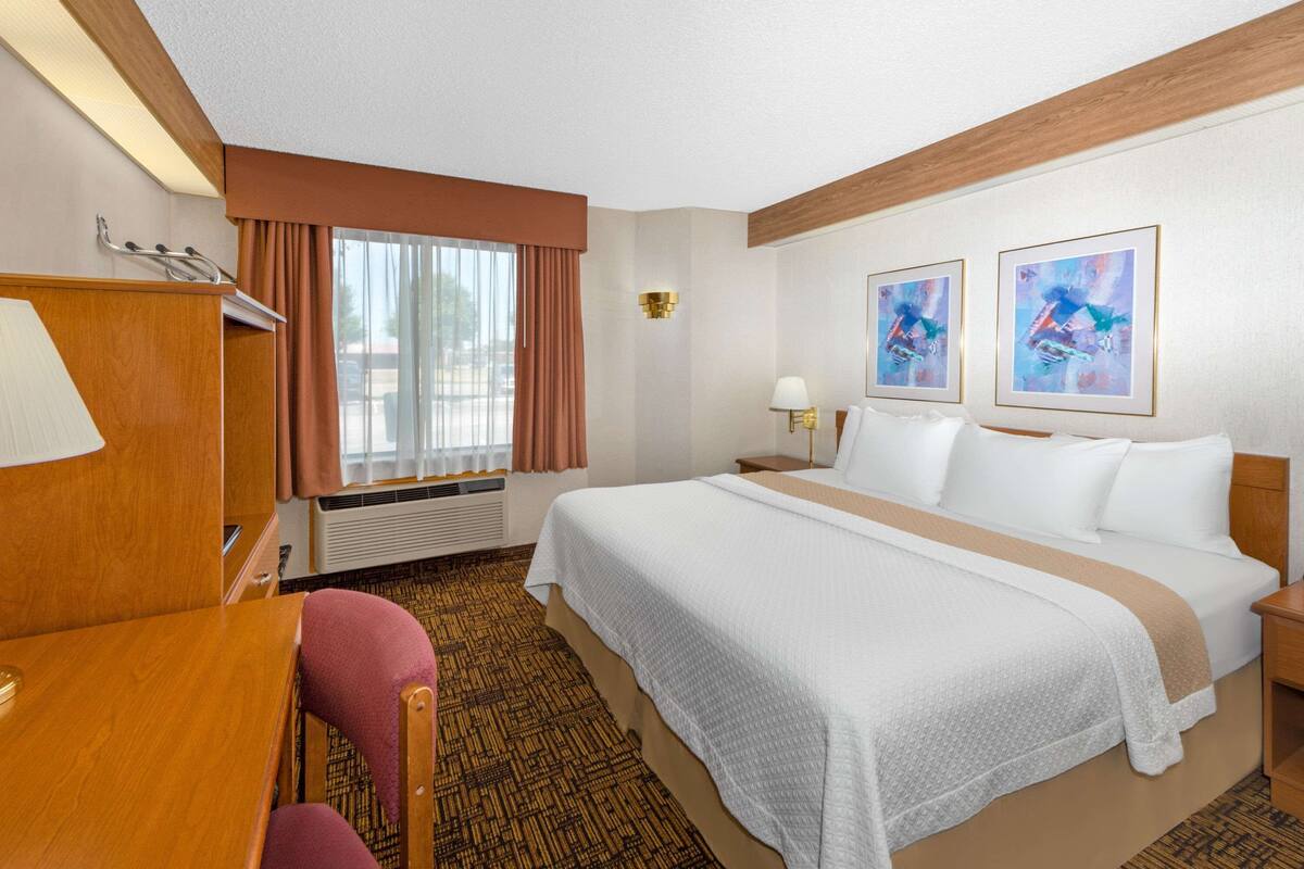 A hotel room with a large bed, a desk, a chair, a window with curtains, a wall-mounted lamp, and two framed artworks.