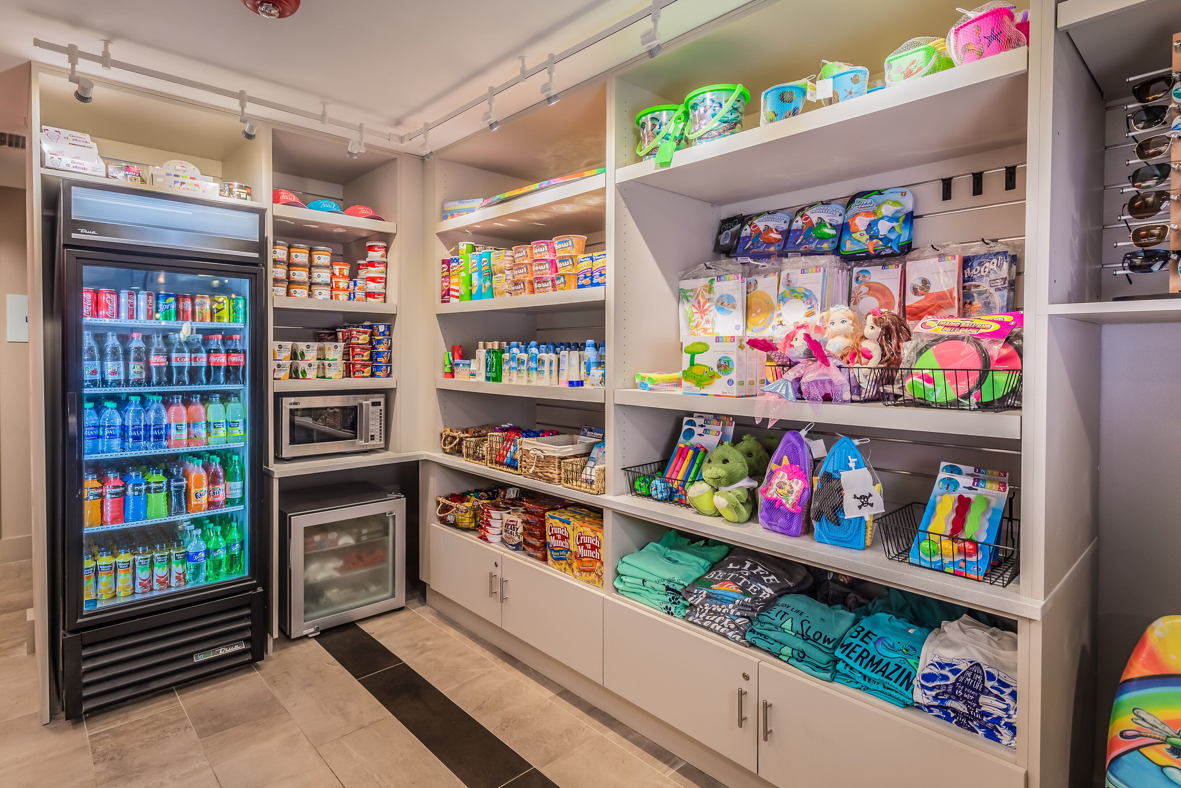 En velorganiseret butik med køledisk til drikkevarer, hylder fyldt med snacks og et udvalg af varer, herunder legetøj og tøj.