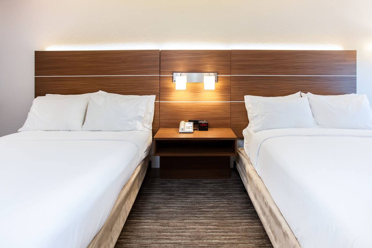 Habitación de hotel con dos camas, cabecero de madera, mesitas de noche con lámparas y una pequeña mesa con un teléfono y un reloj.