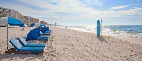 A beach with rows of blue lounge chairs and umbrellas, a surfboard leaning against a pole, and a sandy shoreline with waves.