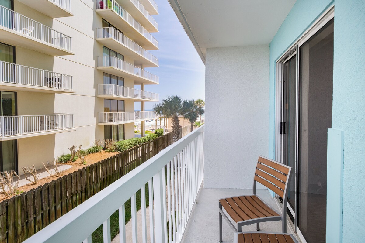 Un balcon avec un banc en bois, une balustrade blanche et une vue sur un immeuble doté de nombreux balcons et de palmiers.