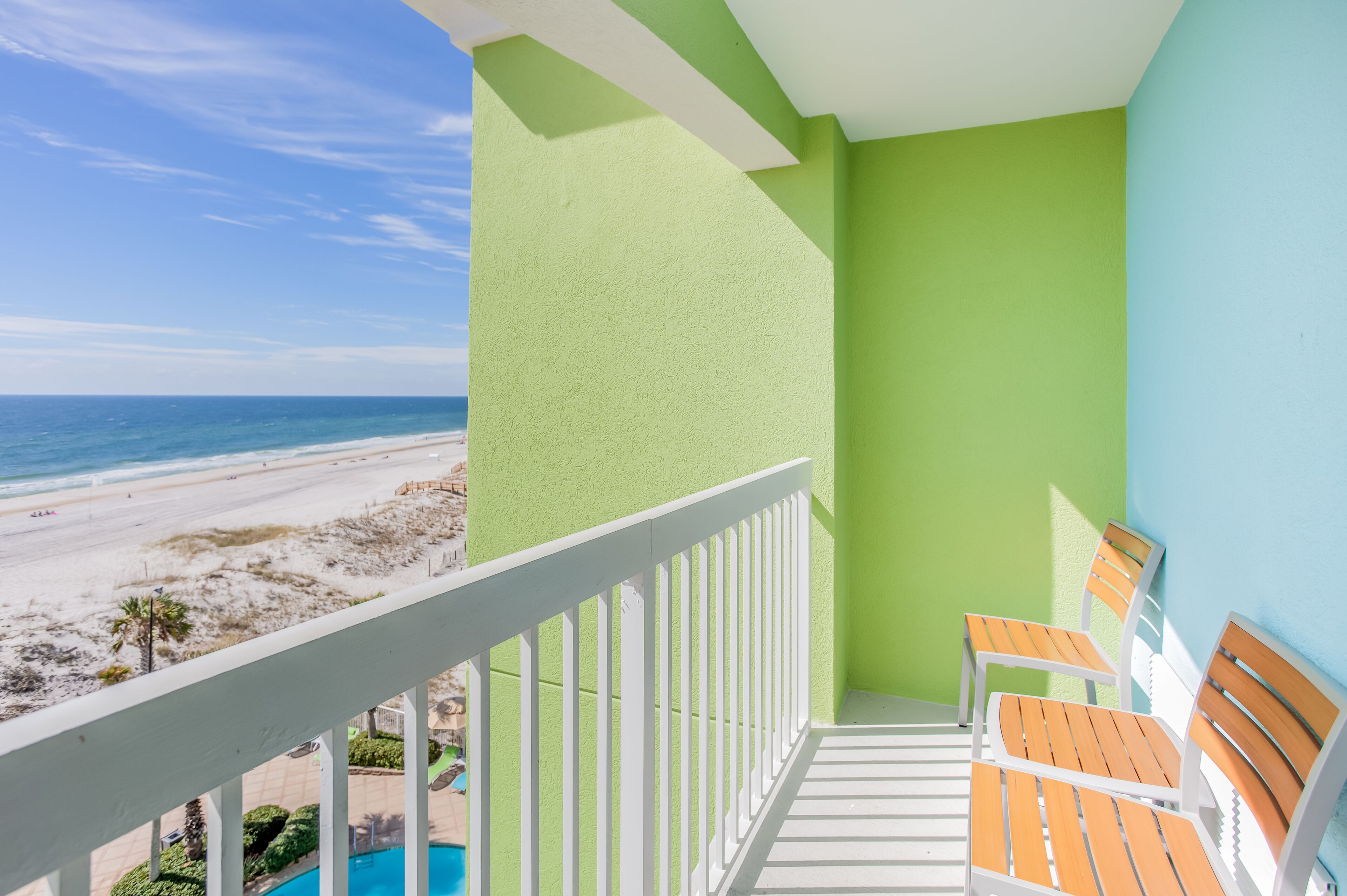 A balcony with a view of the beach, a pool, and two wooden chairs.