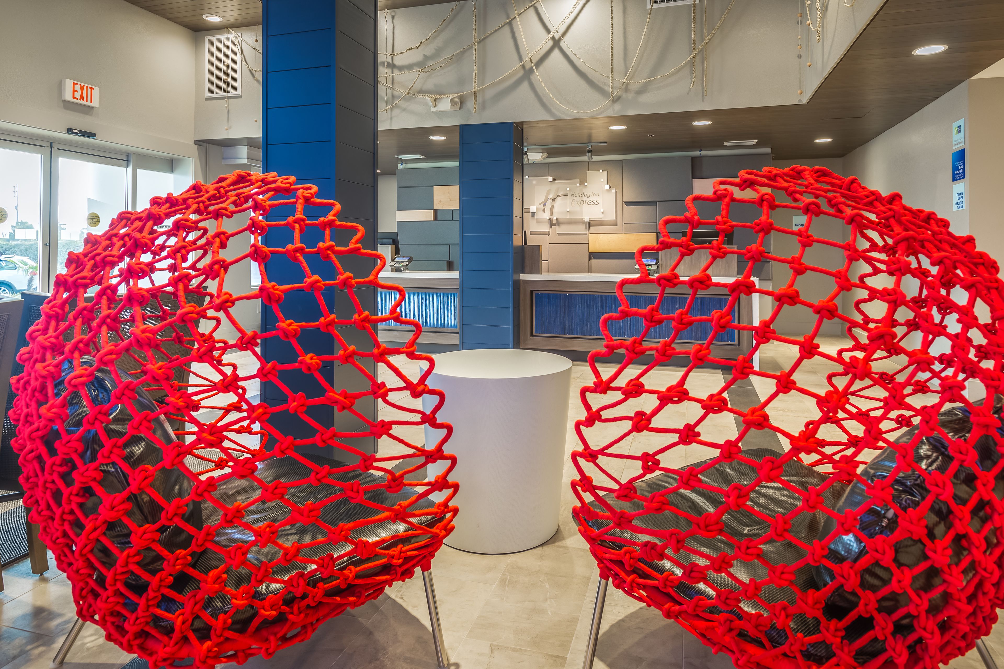 A modern interior with two spherical red chairs with mesh seats and metal legs, a white cylindrical table, and a reception area with a blue accent wall and a sign that reads "EXIT".