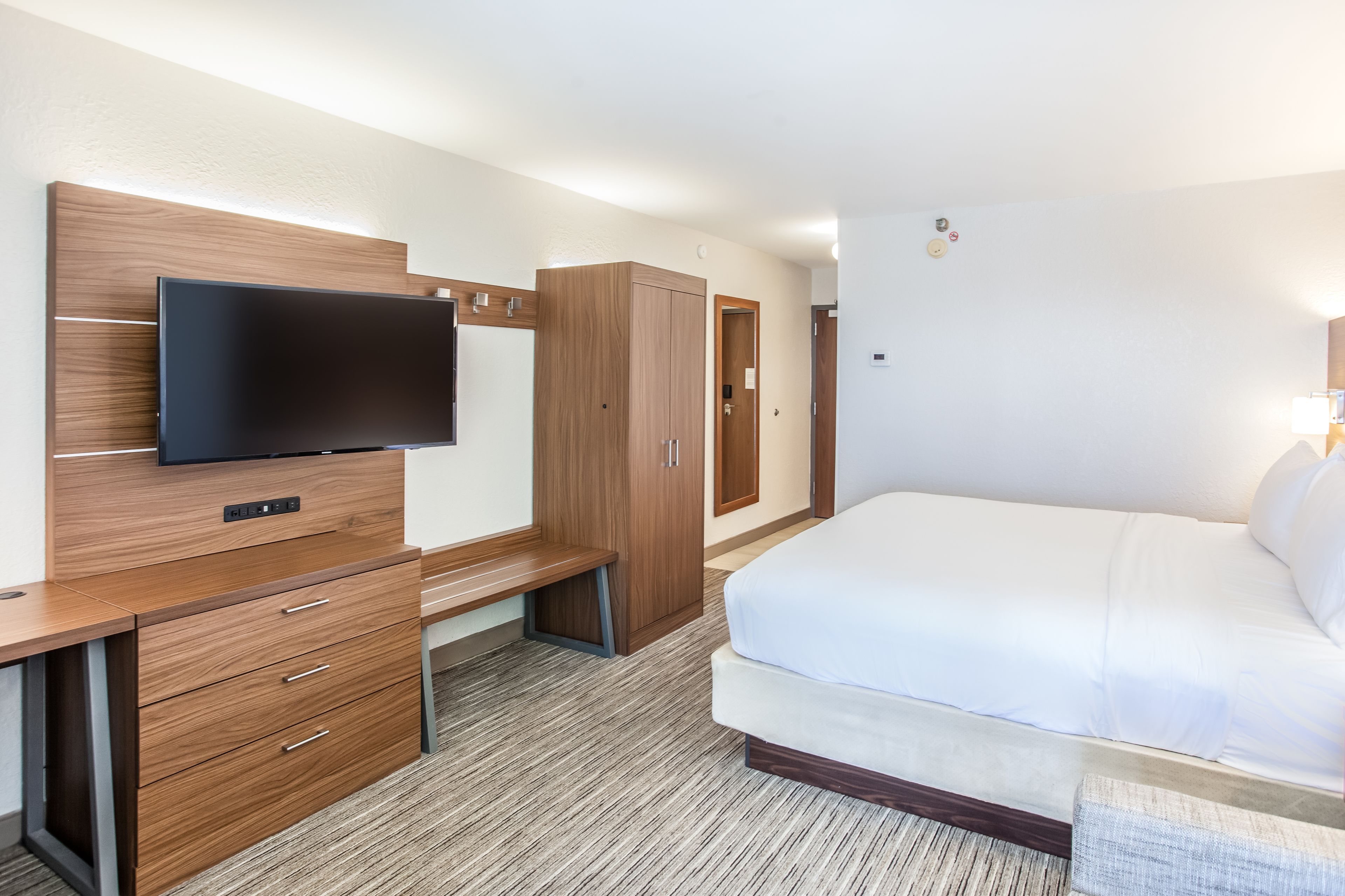 A hotel room with a bed, a desk with drawers, a television mounted on the wall, and a wardrobe.