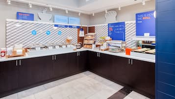 Un área de desayuno con un mostrador que exhibe comida caliente y productos de pastelería, un letrero que anuncia el inicio del día y un letrero para tortitas.