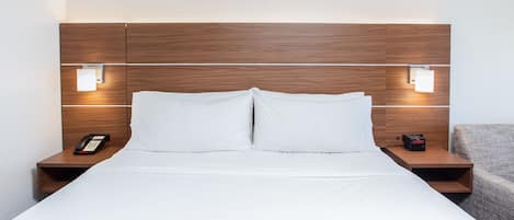 Quarto de hotel com cabeceira de madeira, duas mesas de cabeceira, cama com lençóis brancos e um criado-mudo pequeno com telefone e relógio.