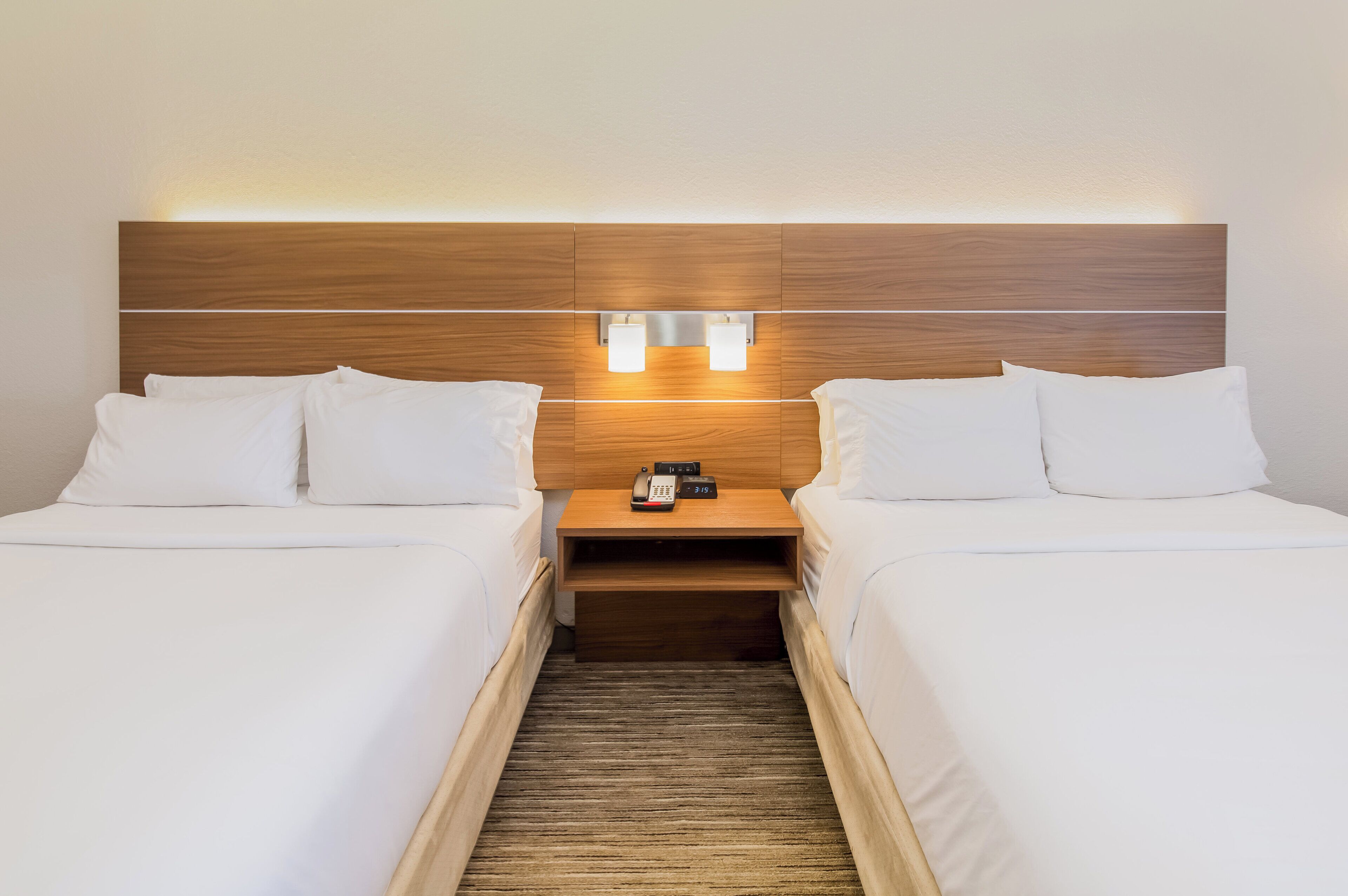 Habitación de hotel con dos camas individuales, cabecera de madera, lámparas de noche, mesita de noche con teléfono y despertador, y suelo alfombrado.