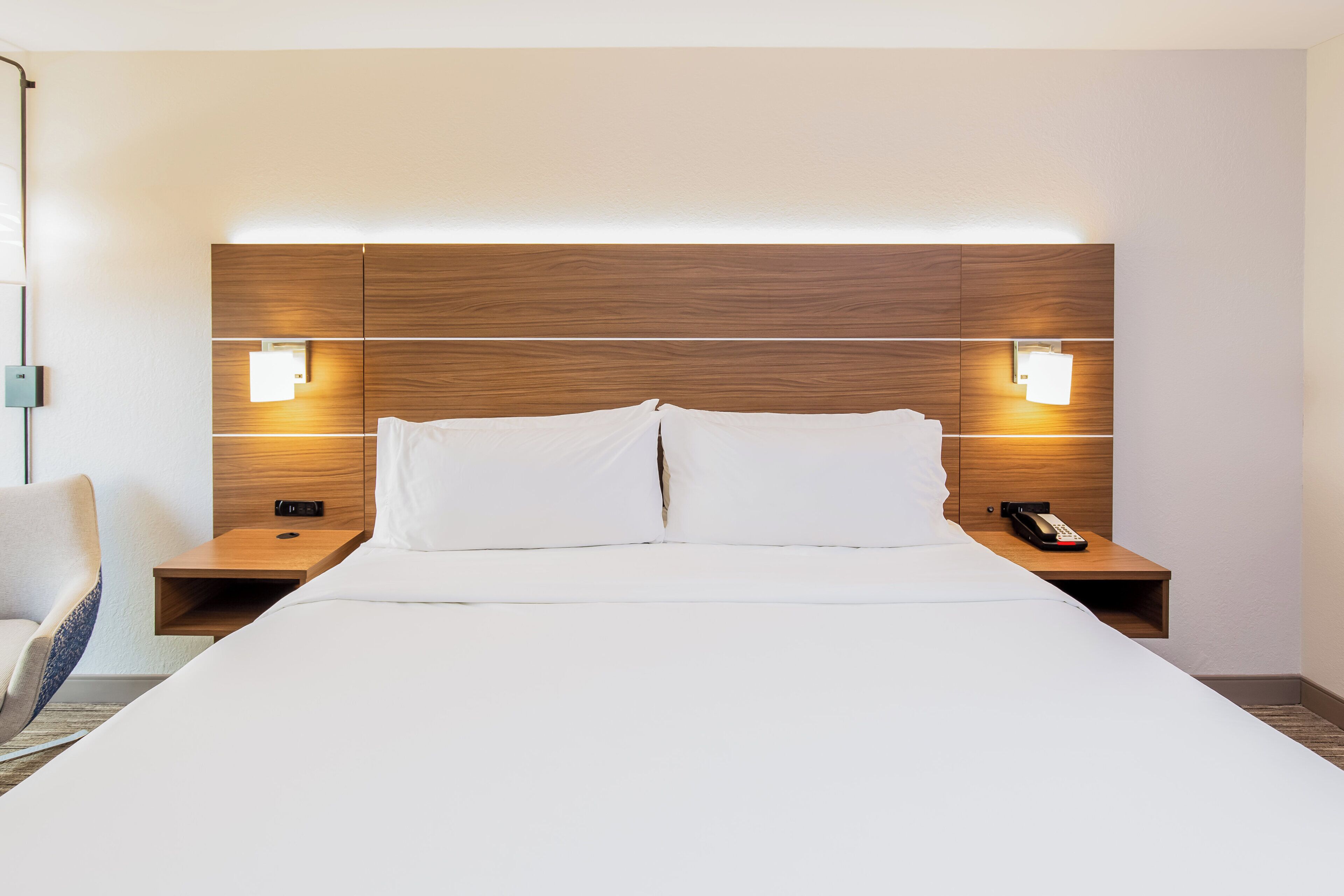 Habitación de hotel con una cama grande, dos mesitas de noche, una silla y un cabecero de madera con iluminación integrada.