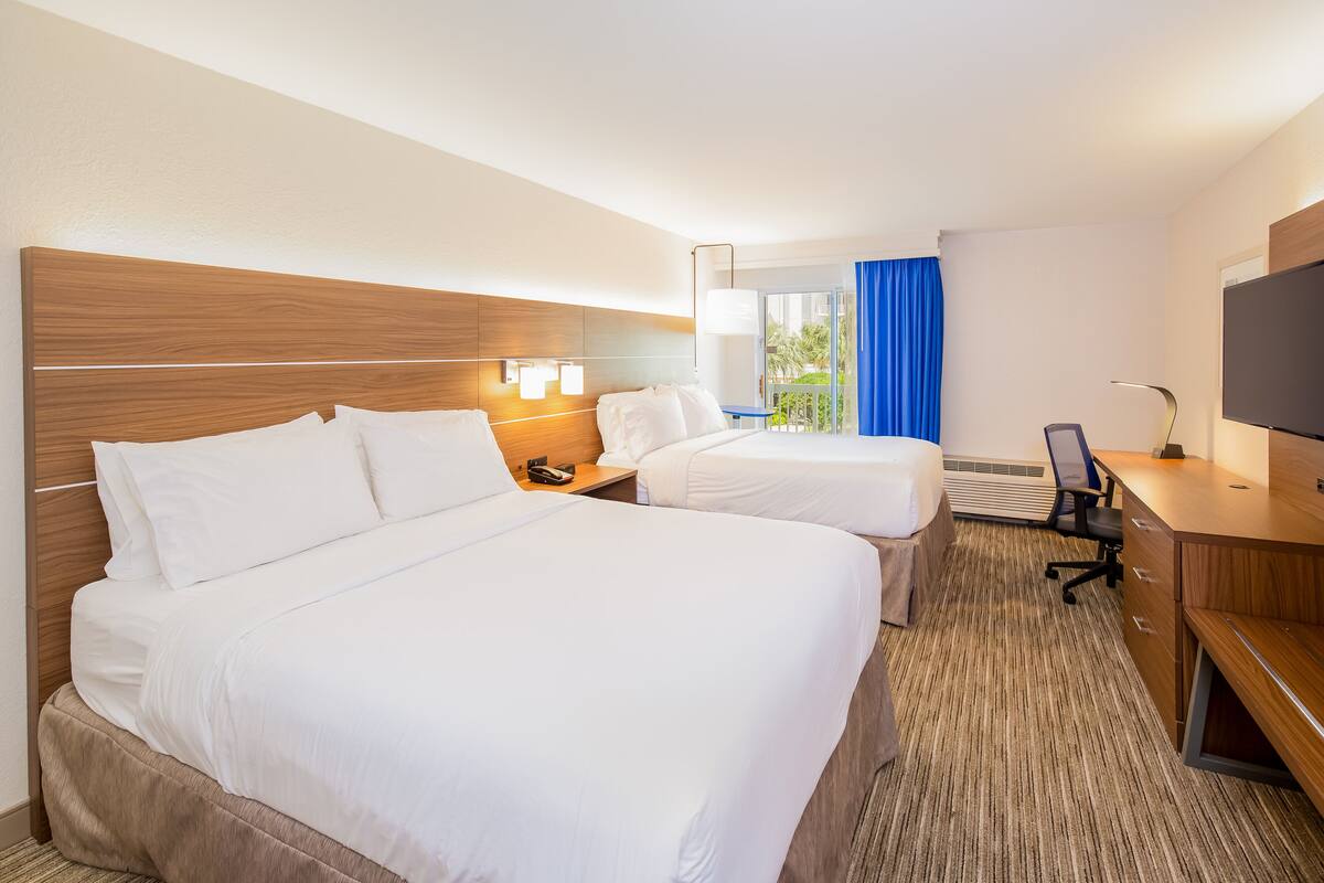 Habitación de hotel con dos camas, un escritorio, una silla, un televisor y una ventana con vistas a zonas verdes.