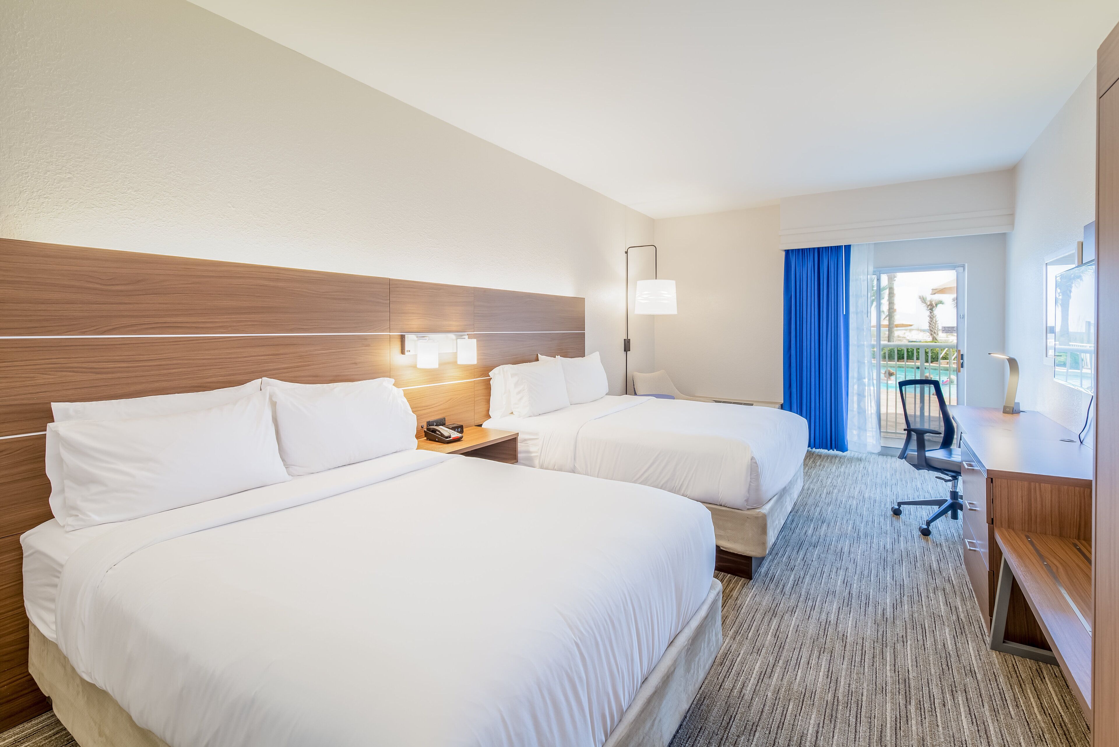 A hotel room with two beds, a desk, a chair, a lamp, and a view of an outdoor area.