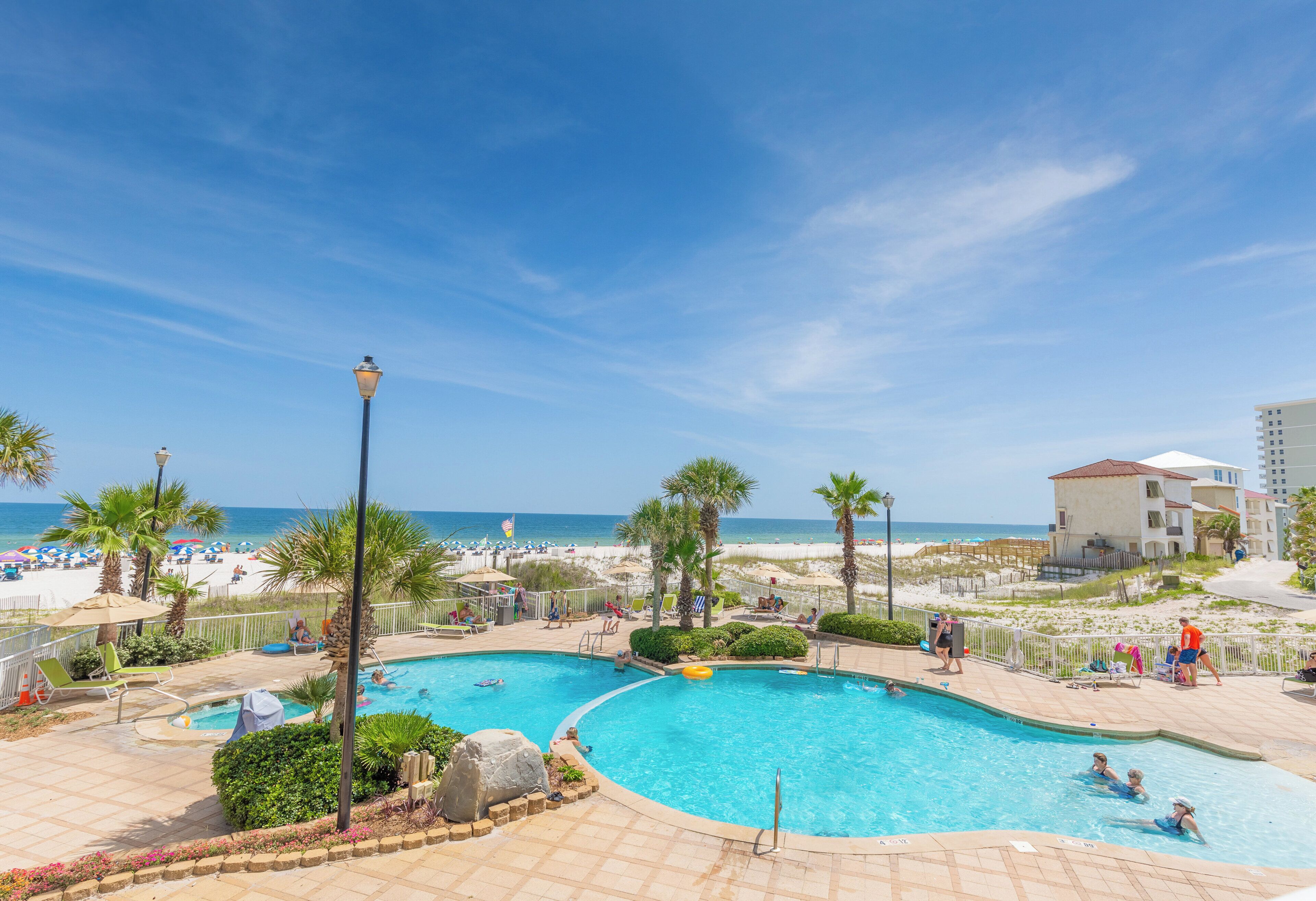 A swimming pool with a curved design, surrounded by lounge chairs and umbrellas, with a beach and ocean in the background.