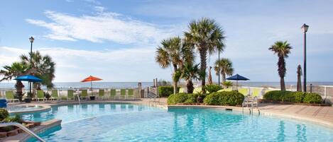 A clear blue pool with a palm tree-lined deck, a view of the ocean, and a beach in the background.