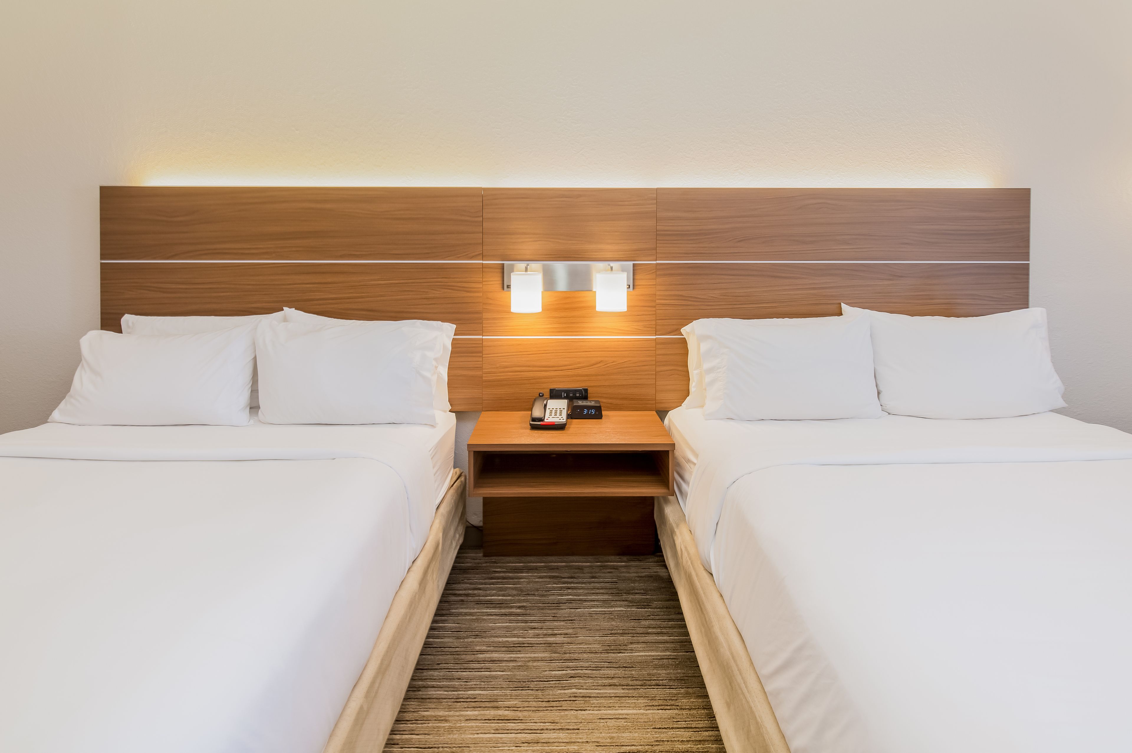 A hotel room with two single beds, a wooden headboard, bedside lamps, a nightstand with a phone and a clock, and a wooden floor.