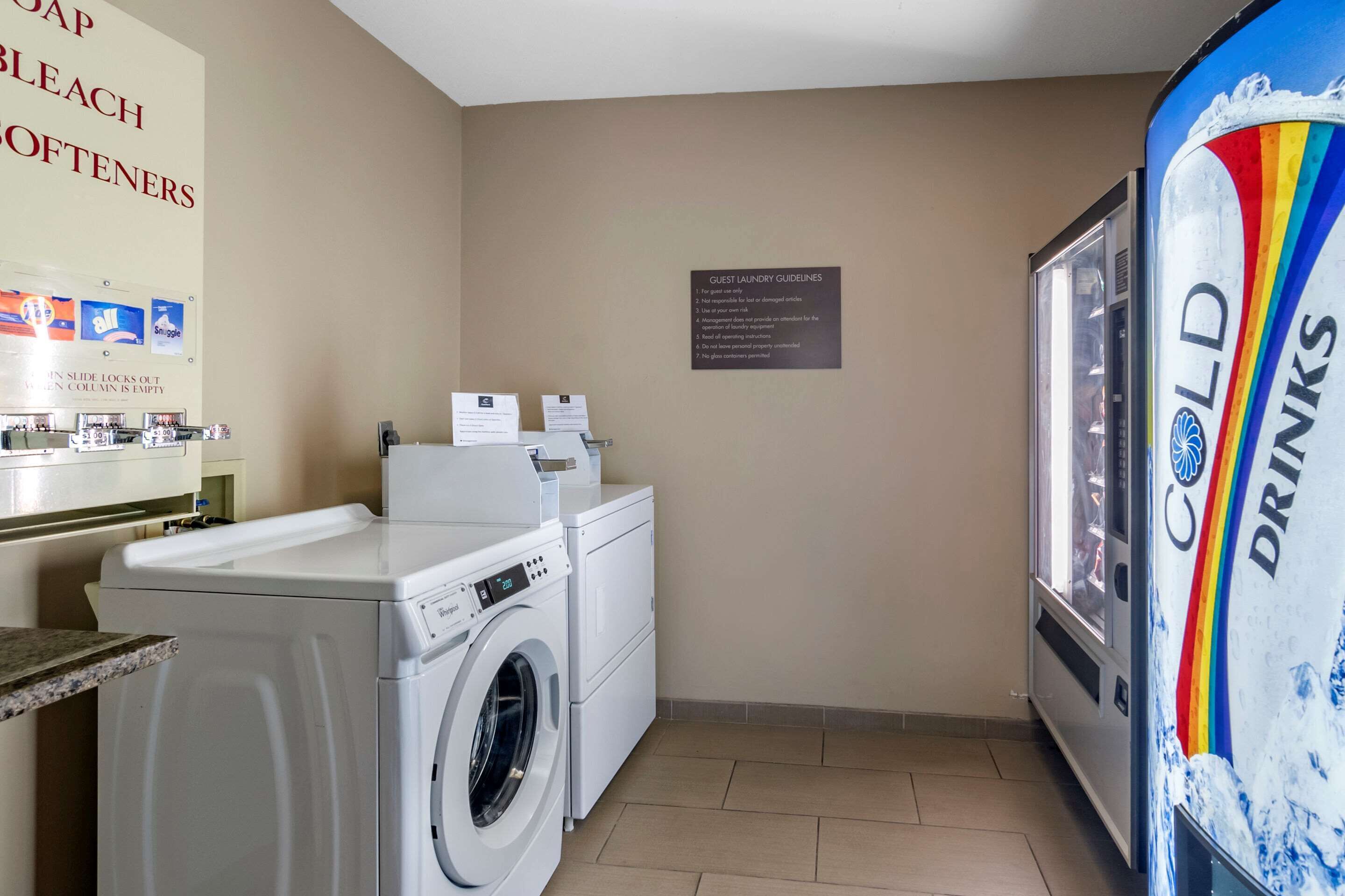 A hotel laundry room with a washing machine, dryer, and a refrigerator. There are signs for guest laundry guidelines and a sign for cold drinks.