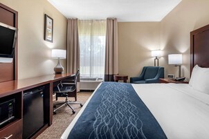 Una habitación de hotel con cama, escritorio, silla, televisor, lámparas y ventana con cortinas.
