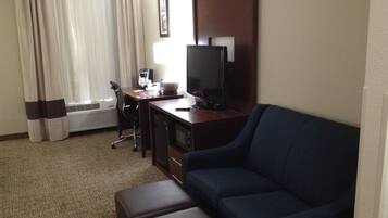 A hotel room with a dark blue sofa, a wooden desk with a television, a coffee maker, and a chair. There is a window with curtains and an air conditioning unit.