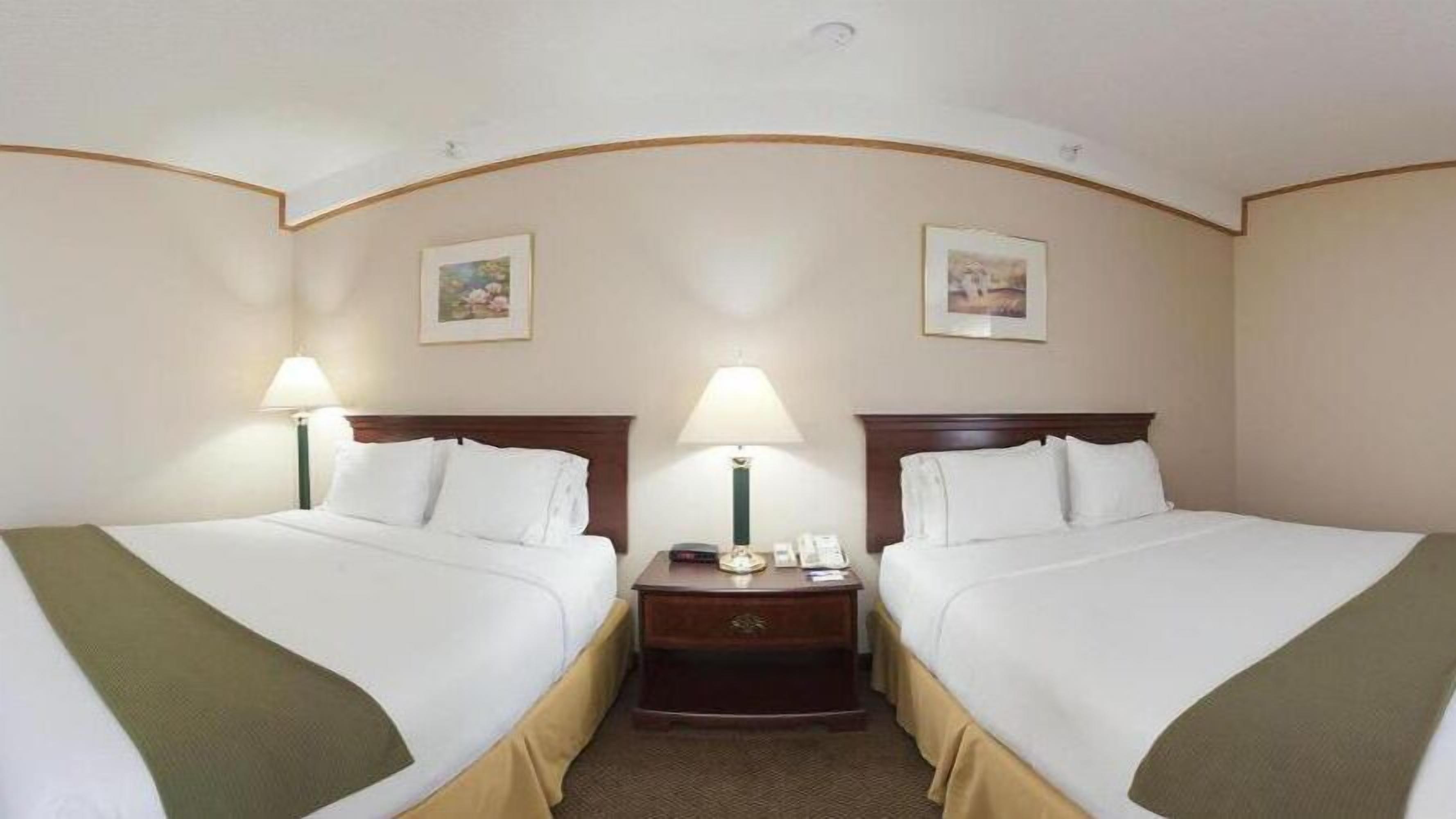 Hotelkamer met twee bedden, elk voorzien van witte lakens en een groene plaid. Twee nachtkastjes met lampen, en de muren zijn versierd met ingelijste foto’s.