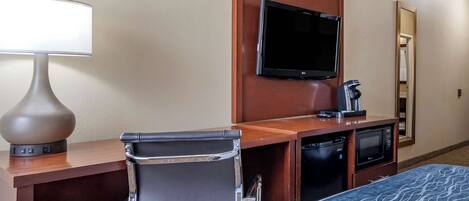 A hotel room with a bed, a desk with a lamp, a flat-screen TV, a microwave, a coffee maker, and a chair.