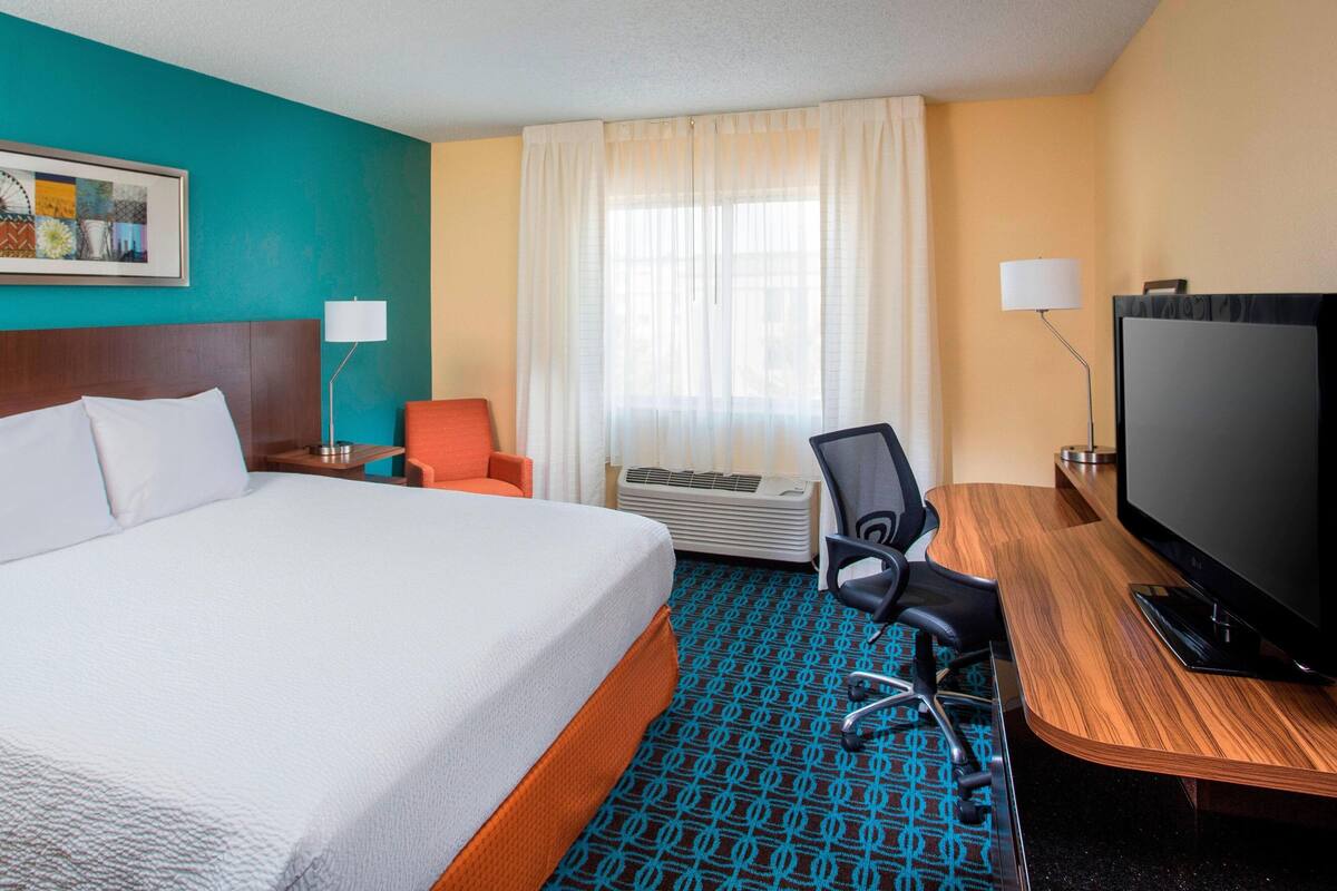 A hotel room with a large bed, a desk with a television, a chair, and a window with curtains.