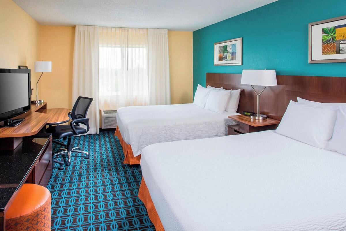 A hotel room with two beds, a desk with a TV, a chair, and a patterned carpet.