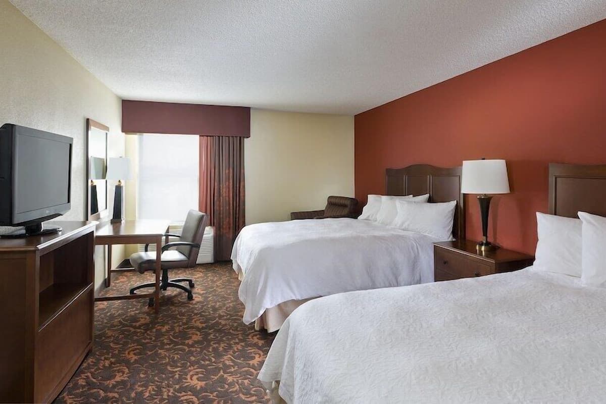A hotel room with two beds, a desk with a television, a chair, and a lamp.