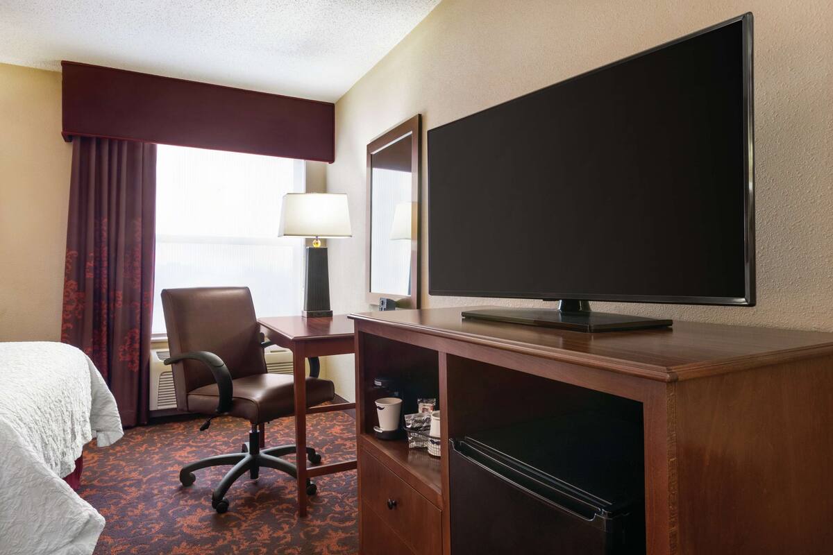 A hotel room with a bed, a desk with a computer, a chair, a television, and a lamp.