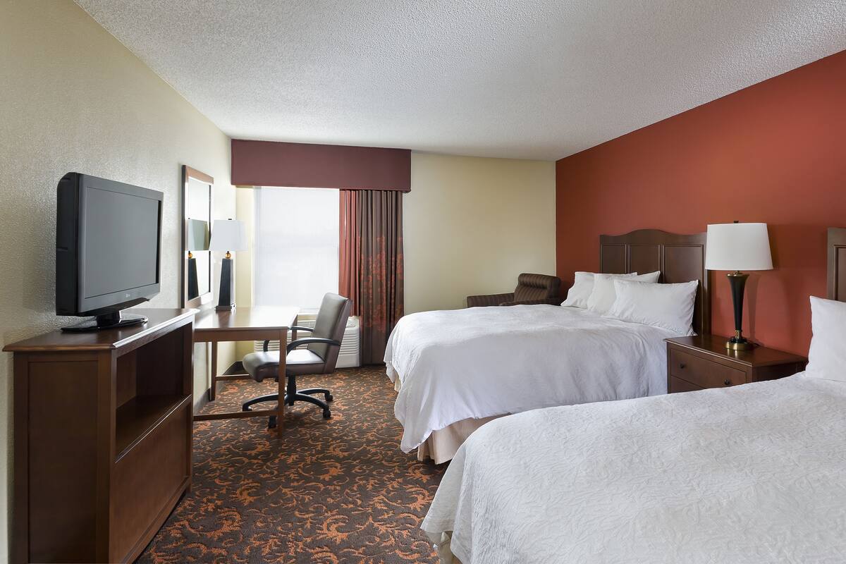 A hotel room with two beds, a desk with a television, a chair, and a lamp.