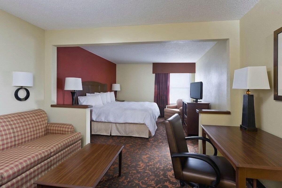 A hotel room with a bed, a sofa, a desk, a chair, a television, and lamps.