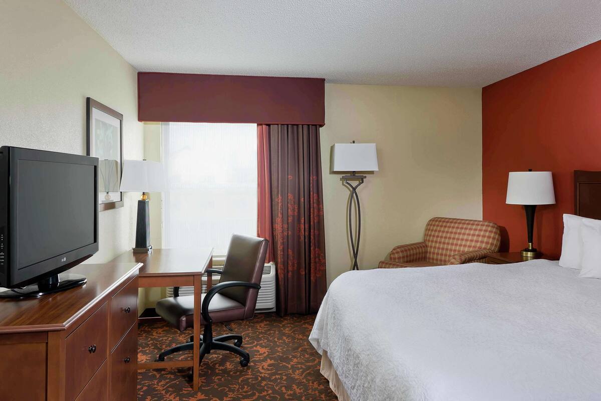 A hotel room with a bed, a desk with a television, a chair, a lamp, and a window with curtains.